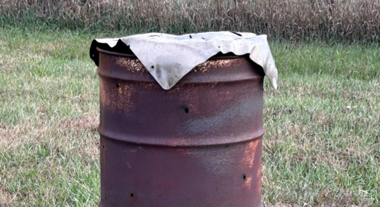 rusty homestead burn barrel with metal cover sitting in a grassy yard
